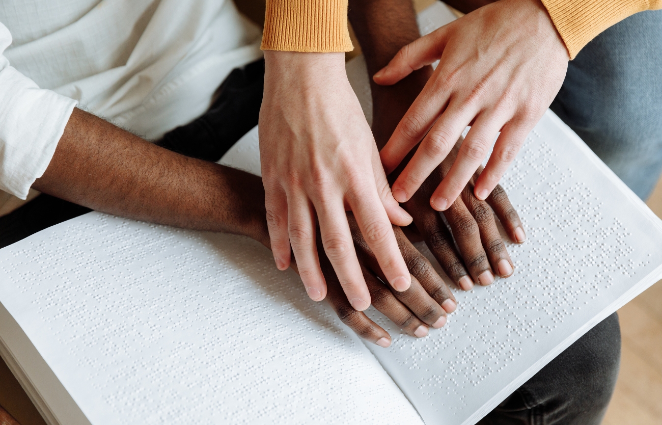 Imagem de uma professora a auxiliar uma pessoa cega na leitura tátil de um livro em Braille