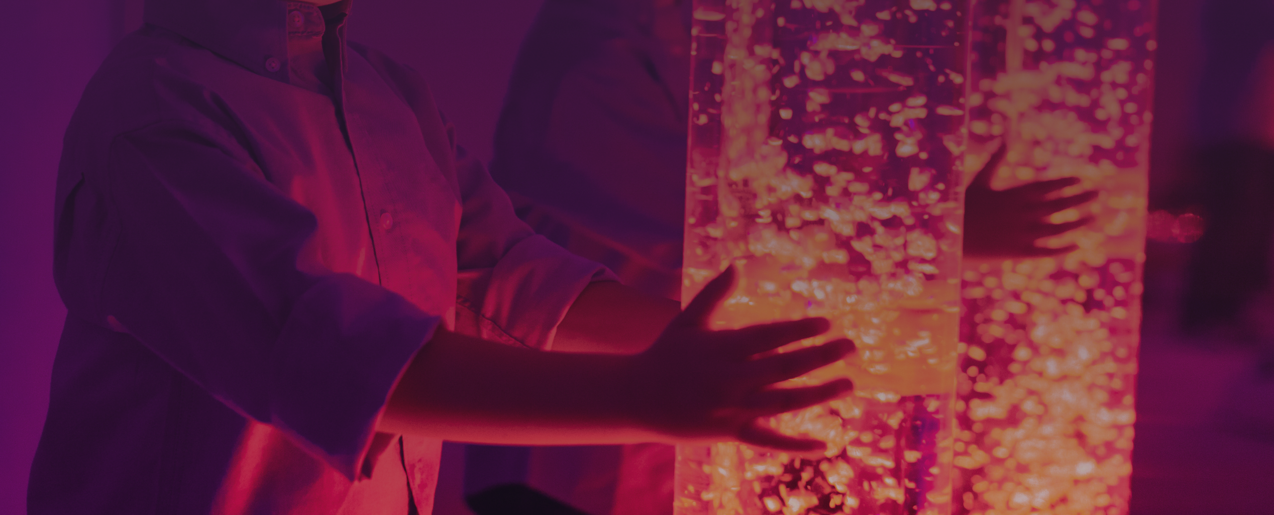 Pessoa a explorar uma coluna sensorial de bolhas iluminada em tons de vermelho, laranja e rosa, num ambiente com iluminação violeta, promovendo a estimulação visual e tátil.